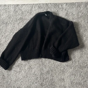 Svart stickad kofta från Zara - Mysig svart stickad kofta från Zara i storlek L. Koftan har en öppen front, breda ribbade muddar och är långärmad. Perfekt för lager-på-lager och chill vibes. Materialet känns fluffigt och ger en avslappnad look.