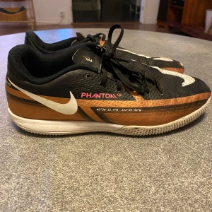 Nike Phantom GT fotbollsskor svart/koppar - Nike Phantom GT fotbollsskor i svart och koppar med vita detaljer och stora Swoosh-loggor. Skorna har snörning, låg profil och platt sula för inomhusspel. Ovandelen är i syntetmaterial med textur för bättre bollkontroll. Snygg och sportig look för dig som älskar fotboll.