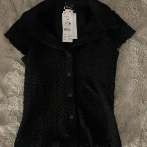 Svart kortärmad blus från Gina Tricot - Svart kortärmad blus från Gina Tricot med snygg struktur i tyget och klassisk krage. Blusen har knappar framtill och en figurnära passform som ger en stilren look. Perfekt att matcha med jeans eller kjol för en trendig outfit.