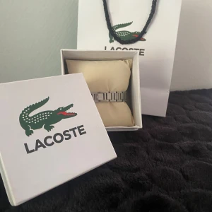 Äkta silver Lacoste armband - Stilren klocka från Lacoste med länkarmband i silverfärgat metall. Klassisk design med Lacoste-loggan ingraverad på spännet. Kommer i originalförpackning och med Lacoste-presentpåse. Perfekt accessoar för dig som gillar en clean och modern look.