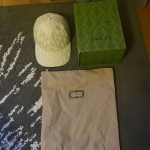 Vit gucci Keps med Box och Dustbag - Snygg Vit keps från Gucci klassiskt GG-monogram över hela kepsen. Kepsen har böjd skärm och justerbar passform. Kommer med original dustbag och grön Gucci-box. Perfekt för dig som vill ha en exklusiv och trendig accessoar.