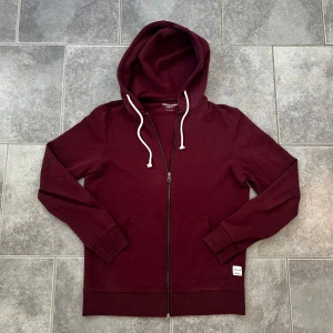 Hoodie Jack and jones  - Säljer en vinröd full zip hoodie ifrån märket Jack and Jones. Tröjan har en lite fläck på magen men ytterst liten. Tröjan har en dragkedja som fungerar klockrent tröjan är knappt. Hör av dig om det skulle vara en minsta fundering. MVH LUNDGRENS