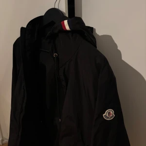 Svart vindjacka från Moncler med huva - Svart ÄKTA vindjacka från Moncler med klassisk logga på ärmen och vit-röd-blå detalj på huvan. Jackan har dragkedja framtill, snörning i huvan och diskreta fickor med tryckknappar. Kan gå ner i pris vid en snabb försäljning 