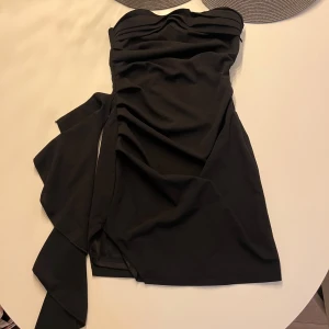 fest klänning  - Snygg svart axelbandslös kortklänning med draperad överdel och markerad midja. Klänningen har en cool slits och ett dekorativt knytband på sidan som ger extra edge. Perfekt för dig som vill sticka ut med en stilren look. Aldrig använd 