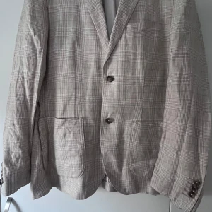 Beige rutig kavaj Slim Fit - Snygg beige kavaj i Slim Fit med rutigt mönster och klassiska knappar på ärmslutet. Tillverkad i ett lätt och luftigt rent linne tyg, perfekt för dig som gillar stilrena och moderna plagg. Kavajen har två framfickor och en slits bak för extra rörelsefrihet.Str 52 och i mycket bra skick.