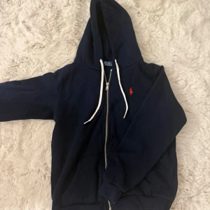 Marinblå hoodie från Polo Ralph Lauren - Marinblå hoodie från Polo Ralph Lauren med vit dragsko och dragkedja framtill. Så bra skick aldrig använd. Säljer pga aldrig avänder. Strl XS/S. Ny pris 2295kr. Priset går att diskutera🙌🩷