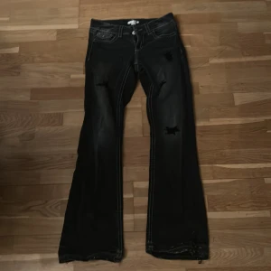 Svarta bootcut jeans med slitningar - Snygga svarta jeans med bootcut passform och slitna detaljer på benen. Jeansen har markerade vita sömmar, klassiska fickor och dragkedja med knapp. Perfekta för dig som gillar en edgy stil och vill sticka ut lite extra.