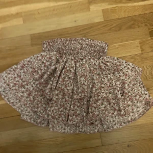 Rosa blommig volangkjol - Supersöt kort kjol i ljusrosa med små vita och gröna blommor. Kjolen har bred smock i midjan och flera volanger som ger en fluffig och lekfull look. Perfekt för vår och sommar när du vill känna dig extra söt och bekväm.