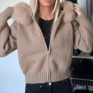 Beige stickad jacka med pälshuva - Säljer en beige stickad jacka med dragkedja och stor fluffig pälskant på huvan. Jackan har ribbade muddar och midja, samt långärmad modell. Perfekt för dig som vill ha en mysig och snygg look under höst och vinter. 