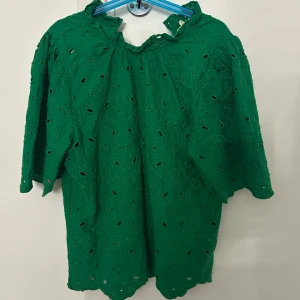 Grön blus med broderade blommor - Supersöt grön blus med broderade blommor och hålmönster. Blusen har korta ärmar, volangkant vid halsen och lätt puff vid axlarna. Perfekt för dig som vill sticka ut med en färgstark och unik topp. Materialet känns som bomull och ger en luftig känsla.