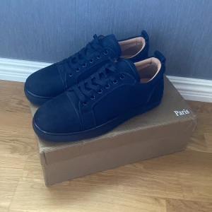 Christian Louboutin svarta sneakers - Snygga svarta sneakers från Christian Louboutin med klassisk röd sula och diskret logga. Skorna har rund tå, snörning och är tillverkade i mocka med läderdetaljer på insidan. Perfekta för dig som vill ha en stilren och lyxig look.