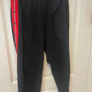 Nike byxor - Svarta mjukisbyxor från Nike med röd och grå rand längs sidan, dekorerad med små Nike-loggor. Byxorna har dragkedjor vid bensluten och elastisk midja. Tillverkade i stretchigt material med 87% polyester och 13% elastan för skön passform. (OBS har bara en ficka, vänster ficka) (om du är intresserad att köpa dom köp dom på Vinted brage2011)