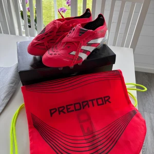 Adidas Predator fotbollsskor röd/vit - Snygga Adidas Predator fotbollsskor i starkt röd med vita och svarta detaljer. Skorna har snörning, klassiska tre ränder på sidan och texten Predator. Ovandelen är i syntetmaterial och sulan har fasta dobbar för gräsplan. Perfekta för dig som vill sticka ut på planen.
