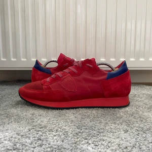 Philippe Model skor - Snygga röda sneakers från Philippe Model med detaljer i mocka och skinn. Skorna har blå accent vid hälen, platt svart sula och klassisk snörning. Skorna har en ikonisk logga på sidan och skorna är i storlek 44! Hoppas du blir nöjd, SBstyle!