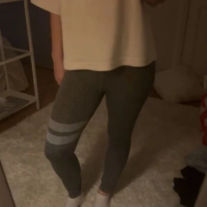 Grå leggings från Stronger - Snygga grå leggings från Stronger med två vita ränder på ena benet. Hög midja med logotyp i midjebandet och tight passform. Perfekta för träning eller chill, stretchigt material som sitter skönt hela dagen.