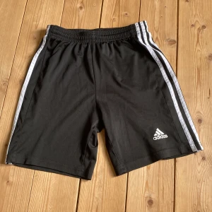 Svarta Adidas shorts med vita ränder - Svarta sportiga shorts från Adidas med klassiska vita ränder längs sidorna och Adidas-logga framtill. Tillverkade i lätt och bekvämt syntetmaterial, perfekt för träning eller chill. Elastisk midja för skön passform. Snygg och enkel design som alltid funkar.