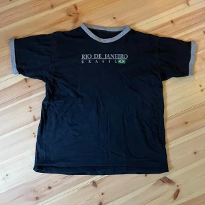 Svart Rio de Janeiro t-shirt - Svart t-shirt med gråa muddar vid ärmslut och halsringning. Tryck på bröstet med texten 'Rio de Janeiro Brasil' och en liten brasiliansk flagga. Klassisk passform och kortärmad, perfekt för en avslappnad stil.