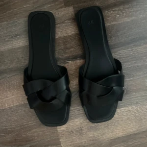 Svarta sandaler med flätad rem H&M - Stilrena svarta sandaler från H&M med fyrkantig tå och breda, flätade remmar över foten. Platt sula och öppen design, perfekta för varma dagar. Snygg minimalistisk look som passar till många outfits.