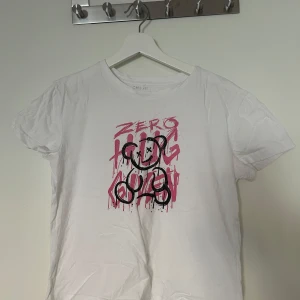 Vit t-shirt med grafiskt tryck - Vit t-shirt med coolt grafiskt tryck framtill i rosa och svart. Trycket föreställer en nalle och texten 'ZERO HUGS' i streetart-stil. Klassisk passform och rund halsringning, perfekt för en edgy vardagslook.