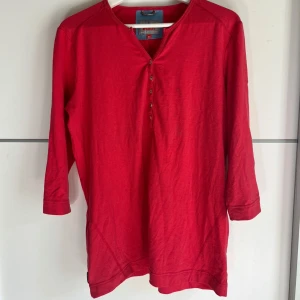 Röd långärmad tröja från McKinley - Röd långärmad tröja från McKinley med diskreta tunna ränder och knappslå framtill. Tröjan har v-ringning och är tillverkad i ett mjukt, lätt material som passar perfekt för utomhusaktiviteter. Broderad logga på ena ärmen ger en sportig touch.