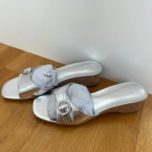 Silvriga sandaler med korkklack från MNG - Glänsande silvriga sandaler från MNG med öppen tå, fyrkantig front och snygg spänndetalj. Skorna har en bekväm korkklack och bred rem i syntetmaterial. Perfekta för att lyfta din sommarstil med en touch av metallic. Helt nya och aldrig använda.
