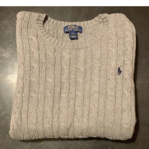 Grå kabelstickad tröja Polo Ralph Lauren - Kabelstickad grå tröja från Polo Ralph Lauren med klassisk rund halsringning och broderad marinblå logga på bröstet. Tröjan är långärmad och har ribbade muddar vid ärmslut och nederkant. Perfekt för lager-på-lager under kyliga dagar.