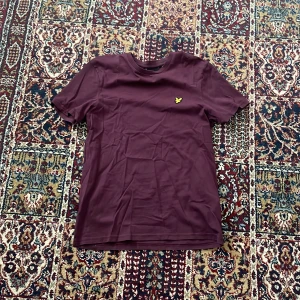 Vinröd t-shirt från Lyle & Scott - Vinröd t-shirt från Lyle & Scott med klassisk passform och korta ärmar. T-shirten har en gul broderad logga på bröstet och är tillverkad i mjuk bomull. Perfekt för en clean och stilren look.