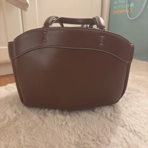 Brun axelväska  - Snygg brun axelväska med kontrasterande vita sömmar och både korta och långa handtag. Väskan har en rymlig insida med en beige innerpåse som stängs med dragkedja. Perfekt för dig som vill ha en stilren och praktisk väska.