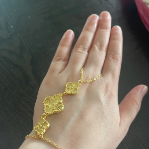 Guldigt blommigt handkedjearmband - Unikt handkedjearmband i guld med tre blommiga fyrklöverformade länkar. Kedjan är tunn och elegant, och de detaljerade blommorna ger en lyxig och trendig känsla. Perfekt för dig som vill sticka ut med något extra snyggt och annorlunda.