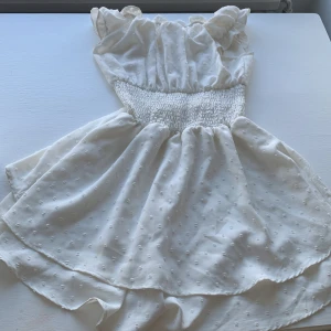 Vit kort sommarklänning med volang - Supersöt vit kortklänning med volangkant och smock i midjan. Klänningen har små diskreta prickar i tyget och en luftig kjol. Perfekt för varma dagar! 