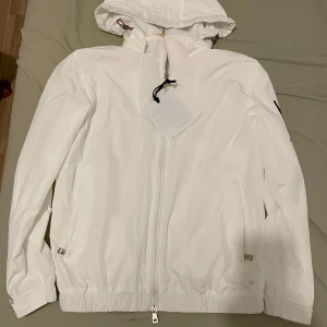 Vit Moncler vindjacka med huva - Snygg vit vindjacka från Moncler med stor svart logga på ryggen och klassisk patch på ärmen. Jackan har huva med dragsko, hel dragkedja framtill och två fickor med dragkedja. Perfekt för dig som vill ha en clean och exklusiv look.