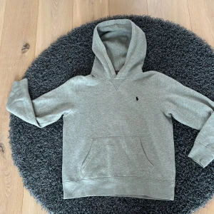 Grå hoodie från Polo Ralph Lauren - Snygg grå hoodie från Polo Ralph Lauren med klassisk broderad logga på bröstet. Tröjan har huva, känguruficka och långa ärmar. Tillverkad i mjuk bomullsmix som är perfekt för chill dagar. Enkel och stilren design som funkar till det mesta.