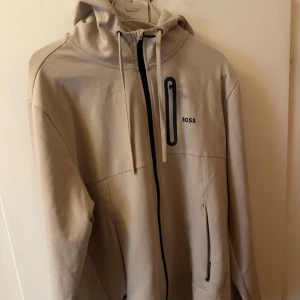 Beige hoodiejacka från BOSS - Snygg beige hoodiejacka från BOSS med dragkedja framtill, huva med snörning och svarta detaljer. Jackan har en bröstficka med dragkedja och två sidofickor. Perfekt för dig som gillar stilren och clean look.
