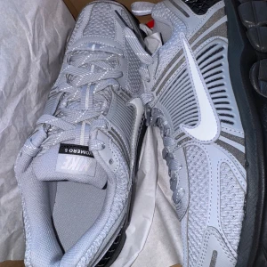 Nike Zoom Vomero 5 grå sneakers - Nike Zoom Vomero 5 sneakers i ljusgrått och svart med vit swoosh. Ovandel i mesh och syntet med coola detaljer och reflekterande partier. Grov sula för extra komfort och snygg urban vibe. Perfekta för dig som gillar techwear och streetstyle. Helt nya original skolåda fås med. Aldrig använda! Storlek 40!