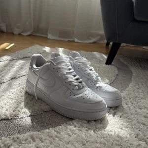 Nike Air Force 1 vita sneakers - Klassiska Nike Air Force 1 sneakers i helvitt skinn med perforerad tå, rund tå och platt sula. Snygg low-top modell med snörning och diskret Nike-logga på sidan och hälen. Perfekta för dig som gillar en clean och tidlös look. Skorna har en liten skada i insida häl som ni kan se på bilden.