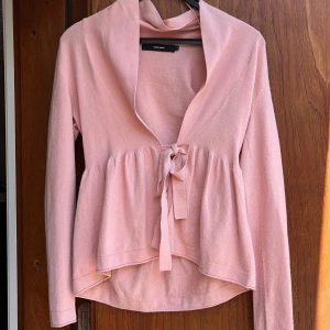 Ljusrosa kofta från Vero Moda - Supersöt ljusrosa kofta från Vero Moda med knyt framtill och volangliknande nederdel. Koftan har lång ärm, bred krage och är gjord i mjukt material som känns skönt mot huden. Perfekt för lager-på-lager och en chill look.