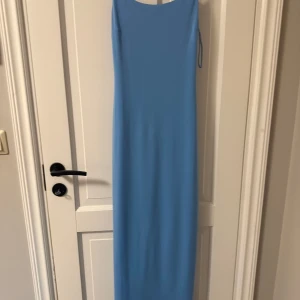 Ljusblå långklänning med smala axelband - Enkel och stilren ljusblå långklänning med smala axelband och rak passform. Klänningen har en minimalistisk look utan mönster eller detaljer, perfekt för dig som gillar clean stil. Materialet ser ut att vara mjukt och följsamt.