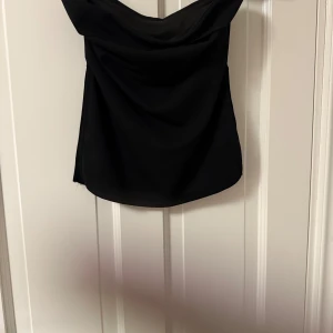 Svart axelbandslös tubtopp - Snygg svart tubtopp med axelbandslös design och lätt draperad framsida. Perfekt för att skapa en stilren och trendig look. Enkel att matcha med både jeans och kjol.