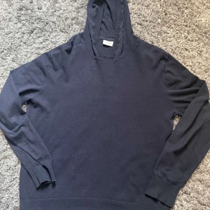 Mörkblå hoodie från Grunt XL - En stilren mörkblå hoodie från Grunt i storlek XL. Tröjan har huva, långa ärmar och är tillverkad i mjuk bomull. Perfekt för en chill och avslappnad look. Ribbstickade muddar vid ärmslut och nederkant ger en snygg finish.