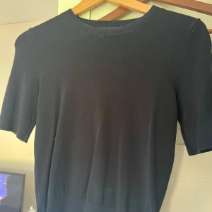 Svart kortärmad tröja från 157 - En stilren svart tröja från 157 med rund halsringning och korta ärmar. Tröjan har ribbade detaljer vid hals och ärmslut, vilket ger en clean och enkel look. Perfekt att matcha med jeans eller kjol för en avslappnad vibe. Typ Oanvänd