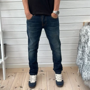 Nudie Jeans blå denimjeans - Säljer ett par klassiska blå jeans från Nudie Jeans modellen Grim Tim med snygga kontrastsömmar och broderad bakficka. Jeansen har normal passform, raka ben och är tillverkade i slitstark bomullsdenim. Perfekta för dig som gillar en tidlös och avslappnad stil.