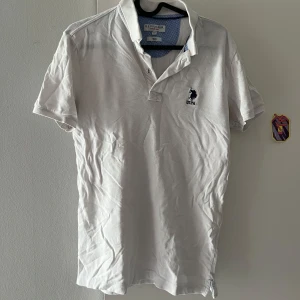 Vit pikétröja från U.S. Polo Assn. - Vit pikétröja från U.S. Polo Assn. i slim fit-modell. Klassisk krage, korta ärmar och broderad blå logga på bröstet. Tillverkad i bomull för en skön och luftig känsla. Perfekt för en clean och sportig stil.