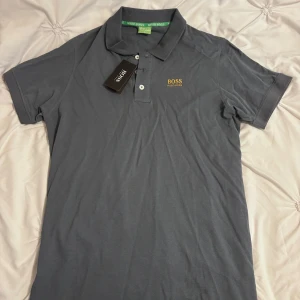 Grå pikétröja från Hugo Boss - Snygg grå pikétröja från Hugo Boss med klassisk krage och knäppning framtill. Tröjan har gul broderad logga på bröstet och gröna detaljer på insidan av kragen. Tillverkad i mjuk bomull, perfekt för en clean och stilren look.