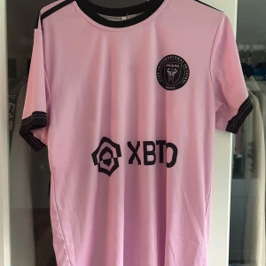 Inter Miami Messi 10 fotbollströja S - Rosa Inter Miami fotbollströja med svarta detaljer och tryck. Messi 10 på ryggen, klubbmärke på bröstet och sponsorlogga framtill. Kortärmad modell i lätt och ventilerande material, perfekt för match eller träning.