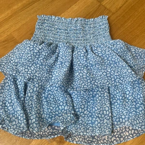  Chelsea kjol - Supersöt blå kortkjol från Made by Chelsea i storlek XS. Klänningen har vitt leopardmönster, smockad överdel och volangig kjol. Perfekt för sommarens alla tillfällen och ger en fräsch och trendig look.