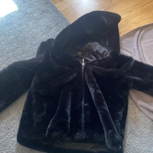 Svart pälsjacka med huva från ONLY - Mysig svart pälsjacka från ONLY i storlek S. Jackan har en fluffig känsla, huva och dragkedja framtill. Perfekt för kalla dagar när du vill vara både varm och snygg. Jackan är helsvart och har en stilren look som passar till det mesta.
