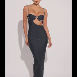 Svart långklänning med cut out - Supersnygg svart långklänning med glittriga axelband och unik cut out-detalj vid midjan. Klänningen har figurnära passform och byst med markerad kupa. Perfekt för dig som vill sticka ut och känna dig extra glam. den är stretchig