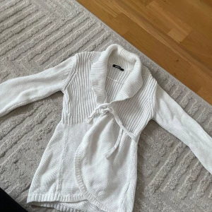 Vit stickad kofta från Gina Tricot - Säljer en vit stickad kofta från Gina Tricot i storlek XS. Koftan har lång ärm, bred krage och knytband i midjan för en snygg omlott-look. Perfekt för lager-på-lager och chill dagar. Supermjuk och enkel att styla till det mesta.