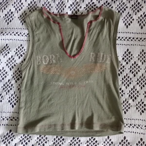 Olivgrön croppad linnetopp Urban Outfitters - Säljer ett supersnyggt olivgrönt linne från Urban Outfitters med tryck 'Born to Ride' och vingar framtill. Toppen har ärmlös design, röd kontrastsöm runt v-ringningen och en avslappnad passform.
