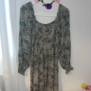 Blommig middlelång med volangärm - Middlelång klänning med blommigt mönster i grönt, lila och beige. Klänningen har långa ärmar med volangdetalj vid ärmslut. Luftigt material, perfekt för vår och sommar.